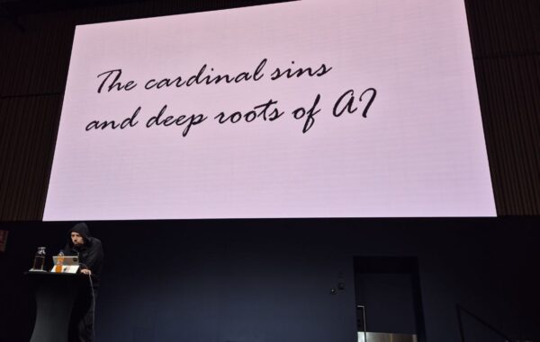 Jouni Heikniemi at the lectern under his closing keynote title slide, The cardinal sins and deep roots of AI, HELish Summit 2026